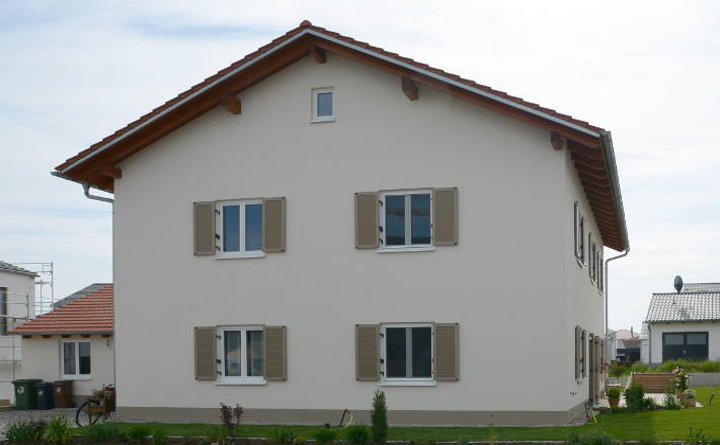 Bild eines neugebauten verputzten Holzhauses. Hausseite mit vier fenstern und beigefarbenen Fensterläden