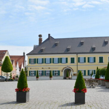 Ansicht des Marktplatzes von Geisenfeld. Im Hinteren Teil ist das Klosterbräu zu sehen. Ein großes gelbes Gebäude mit Torbogen und grünen Fensterläden. Im Vordergrund 3 Säulentujen in Töpfen mit kontrastreicher roter UNterpflanzung. Kopfsteinpflaster