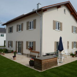 Bild eines weiß verputzten Einfamilienholzhauses mit beigen Fensterläden und Garten mit grünem Rasen