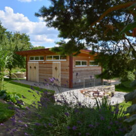 Ein wunderschönes Gartenhaus aus Holz mit 3 Türen und schmalen Fenstern umrahmt von Lavendel, Kiefern und Natursteinen