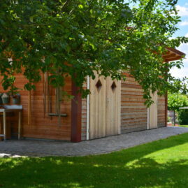 Seitenansicht des Gartenhauses. Gartenwerkzeug hängt geordnet an der Wand. Ein Apfelbaum rahmt das Gartenhaus ein.